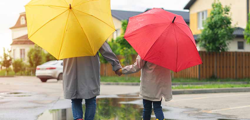 Two people holding hands under yellow and red umbrellas walking on a wet suburban street - https://www.sartori-insurance.com/